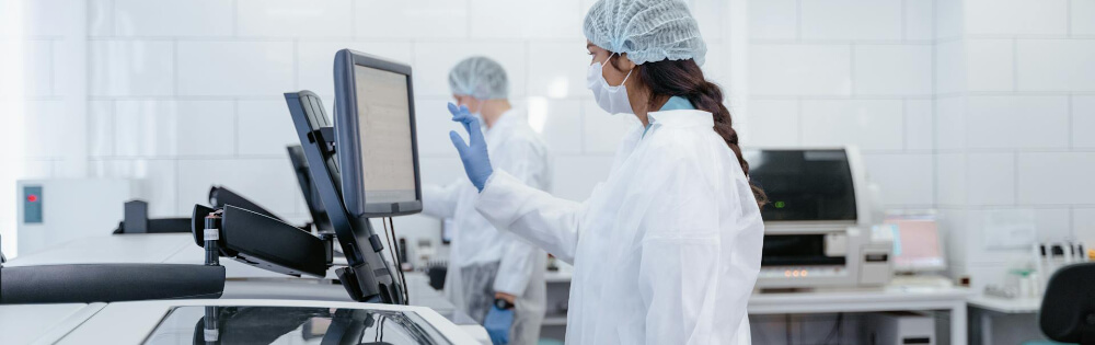 Zwei Laborfachkräfte tragen Schutzkleidung, einschließlich Laborkittel, Haarnetz und medizinischer Masken, während sie an einem Computerbildschirm arbeiten. Sie befinden sich in einem modernen Labor mit verschiedenen Geräten.